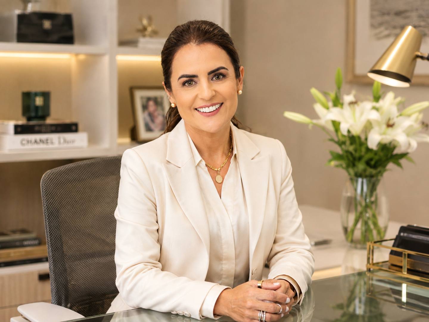 Luciana Xavier sorrindo, de blazer claro, sentada à mesa do escritório